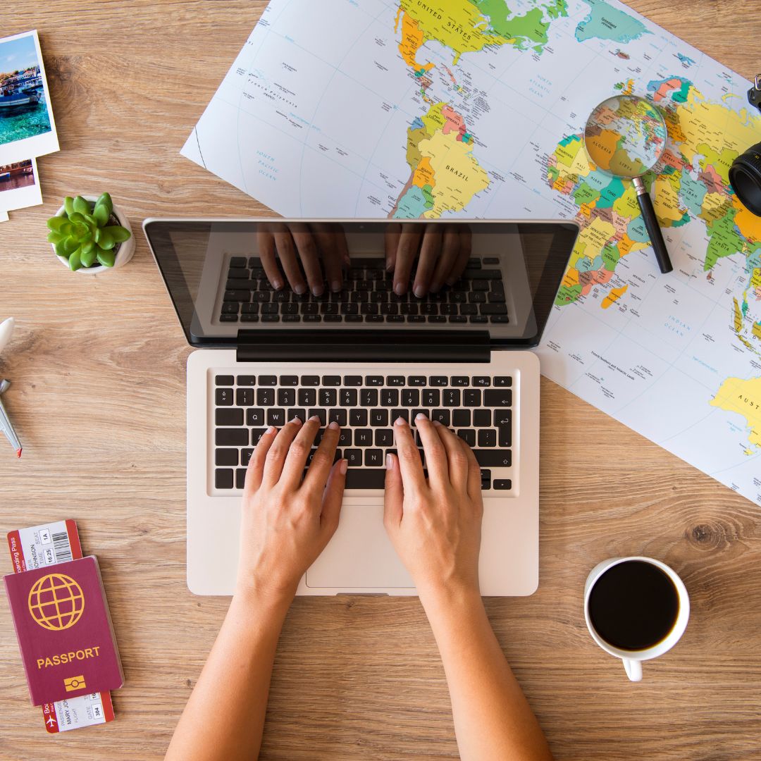 Mani che digitano su un portatile accanto a una mappa del mondo, un passaporto e una tazza di caffè