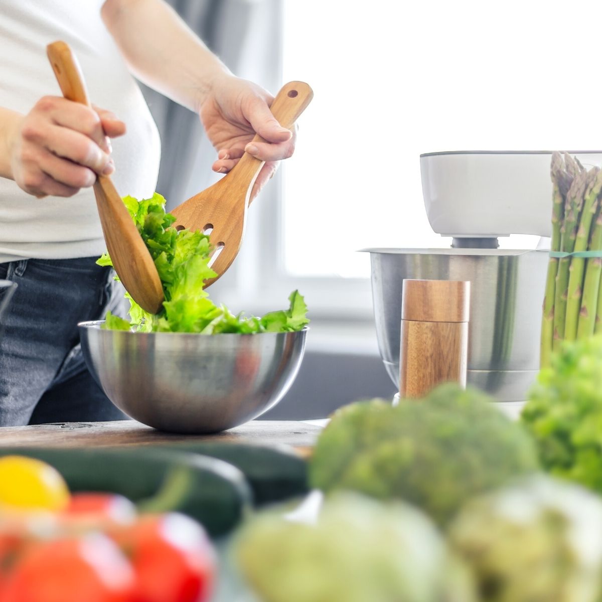 Persona che mescola l'insalata in una ciotola d'acciaio con posate di legno, tra asparagi e verdure fresche