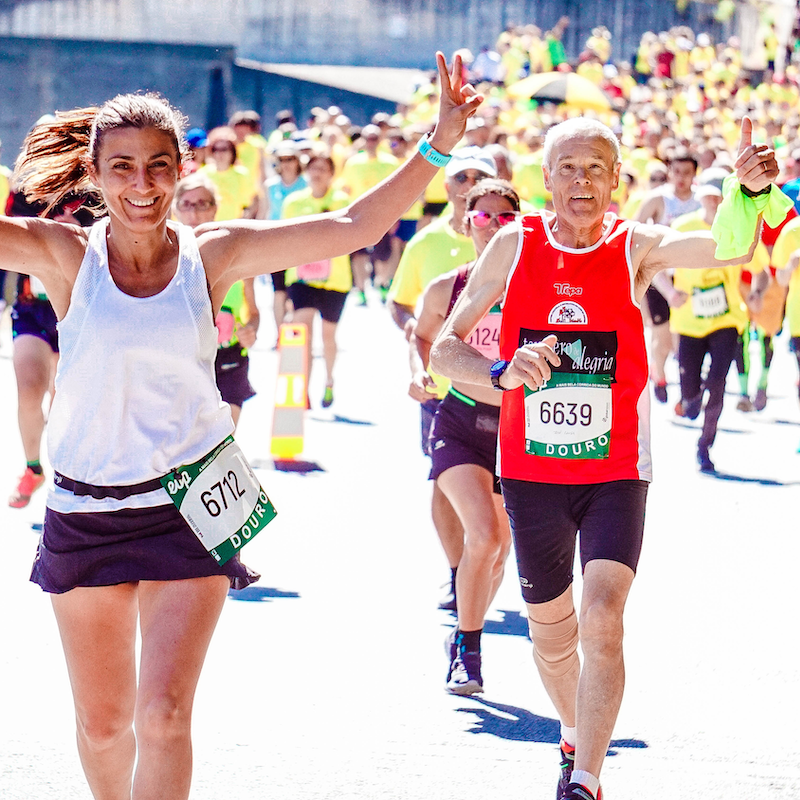 Una donna e un uomo anziano sorridenti con il pettorale durante una maratona, tra molti altri corridori