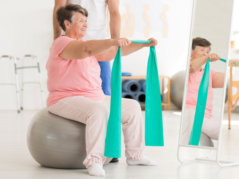 Donna anziana seduta su una palla da ginnastica che tende un elastico, assistita da un fisioterapista
