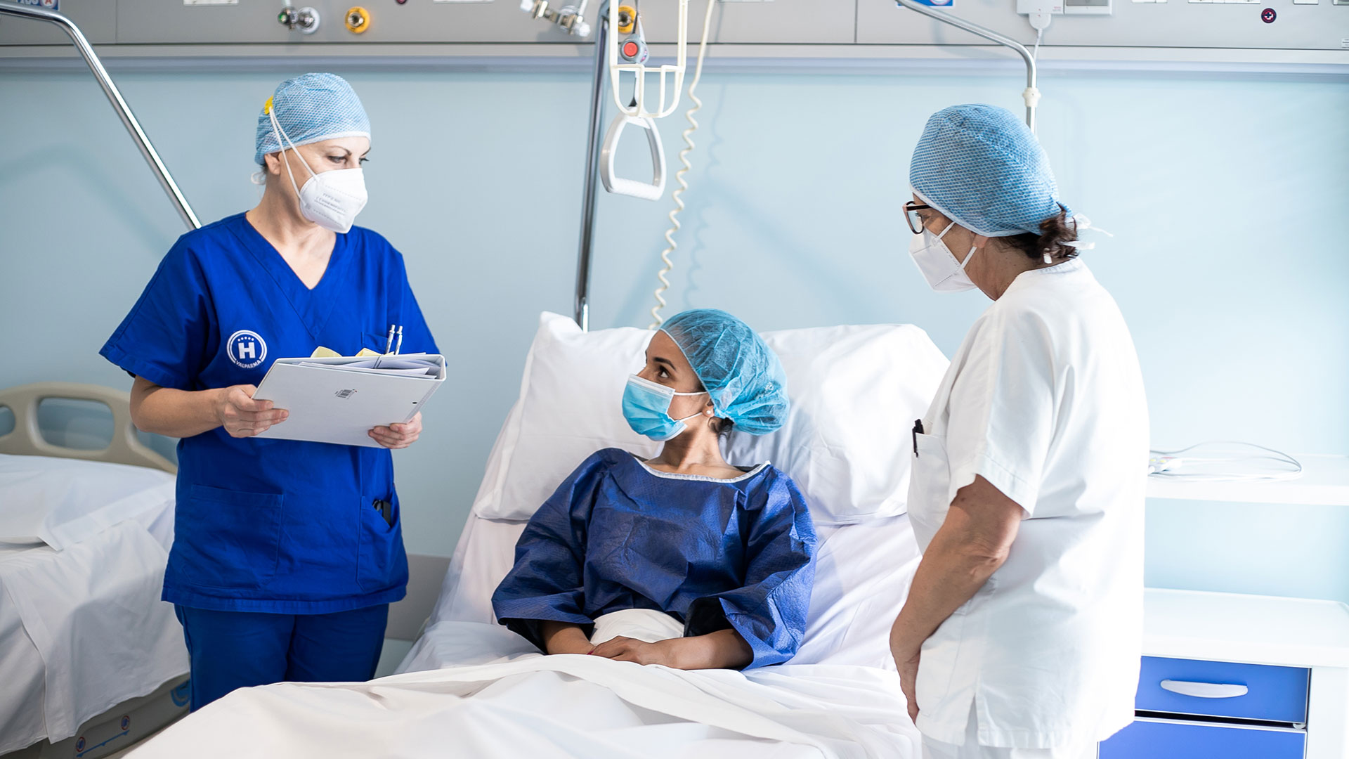Due operatrici di Valparma Hospital parlano con una paziente in cuffia seduta sul letto di degenza