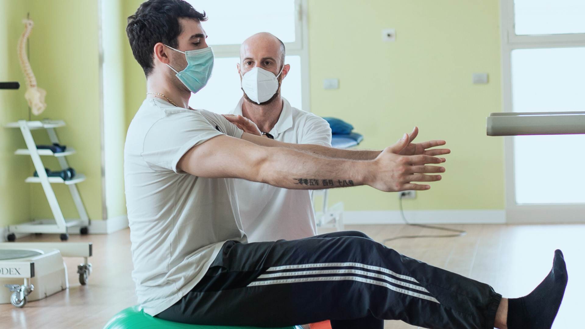 Paziente seduto su una palla verde con braccia tese, seguito da un fisioterapista in una palestra di riabilitazione